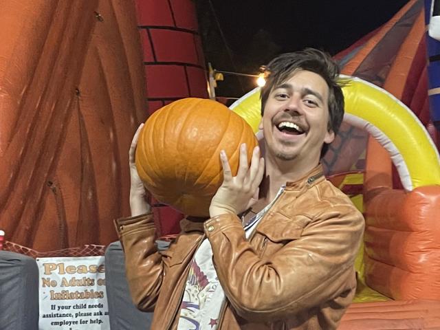 Man in a brown jacket smiling while holding a pumpkin near a colorful inflatable backdrop.